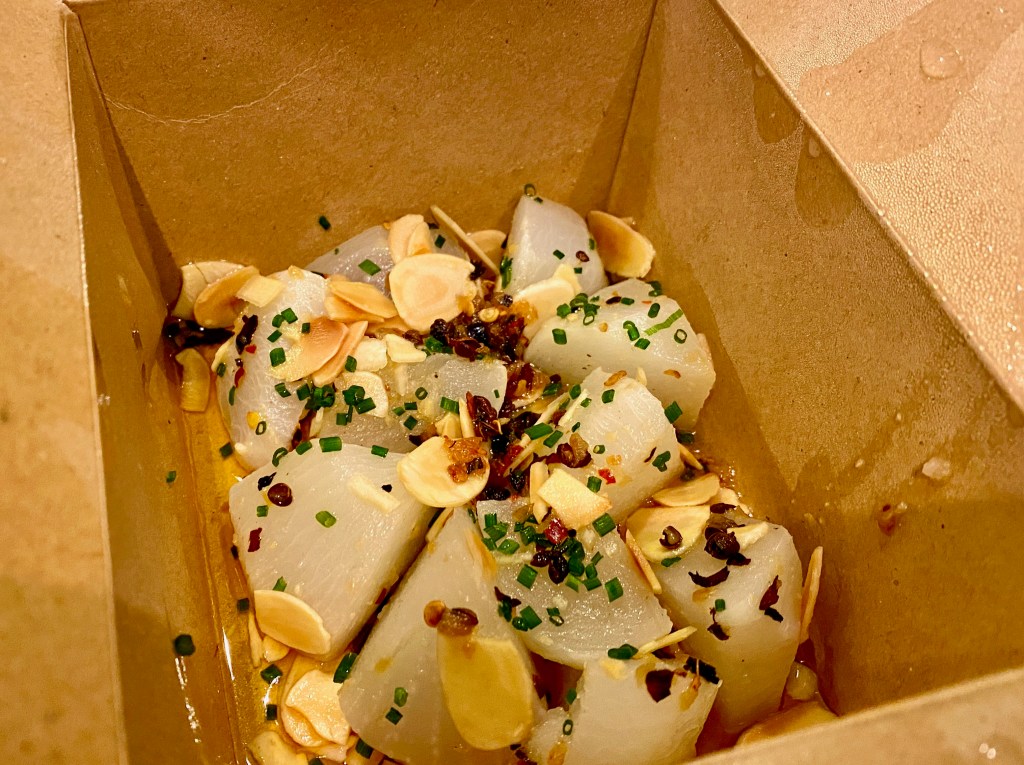 Takeaway carton showing white turnip pieces dressed with slices of almond red crushed Szechuan peppers and chopped chives